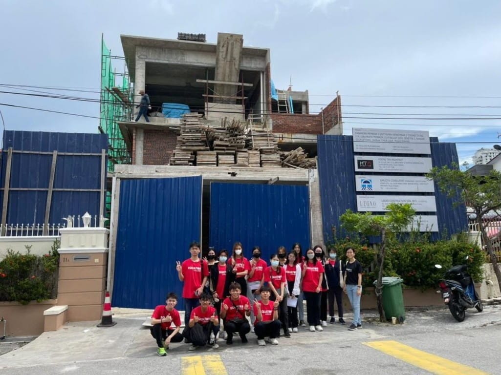 Group poses at construction site with building, blue fence, and sign.